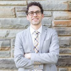 headshot of Dr. Nicholas Reed wearing glasses and a gray blazer with arms crossed leaning against a stone wall