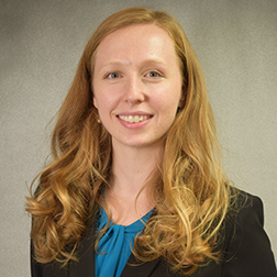 Woman with long, curled strawberry blonde hair, black blazer, and blue blouse. Smiling against a grey background.