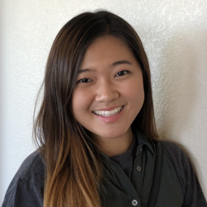 Young woman with long brown hair that becomes more blonde at the tips. She smiles widely and wears a green button up.