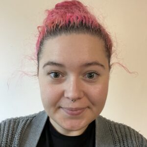 Young woman with pink hair pulled up. She smiles, wearing a grey cardigan and black top.