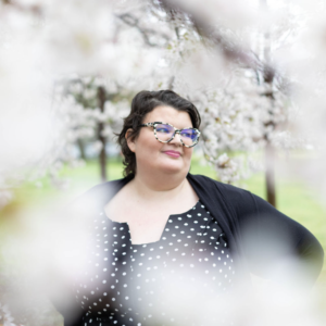 Looking through cherry trees, a woman with short brown hair and tortishell cat-eye glasses is visible, who turns to the right with a black polka dot dress, black cardigan, and hands on her hips.